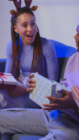 Vertical-Video-Studio-Shot-Of-Gen-Z-Friends-Giving-And-Opening-Presents-For-Christmas-Sitting-On-Sofa-Wearing-Santa-Hat-And-Reindeer-Antlers-3